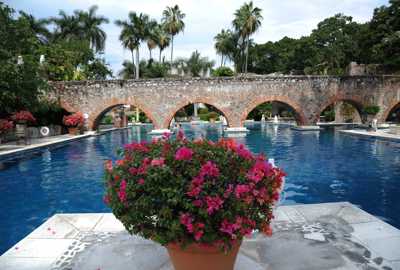 Charming Mexican hacienda with a serene pool, stone arches, and vibrant flowers in a tranquil garden setting. 